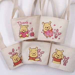 Puede incluir: Seis bolsas de lona con temática de Winnie de Pooh con diferentes diseños. Cada bolsa presenta a Winnie de Pooh y Piglet con diferentes expresiones y accesorios. Las bolsas tienen un fondo crema con detalles en rosa. El texto "Thank u" está impreso en una de las bolsas.