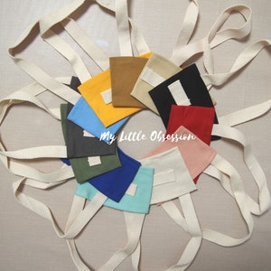 Op de afbeelding: Een verzameling van tien kleine stoffen tasjes met een witte band. De tasjes zijn in verschillende kleuren, waaronder geel, blauw, groen, rood, roze, bruin en zwart. De tasjes zijn in een cirkel op een licht beige oppervlak gerangschikt. De tekst "My Little Obsession" is zichtbaar in het midden van de afbeelding.