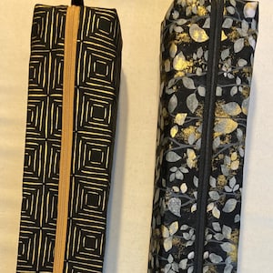 May include: Two long, rectangular zippered cases. The left case is black with a gold geometric square pattern and a gold zipper. The right case is black with a grey and gold leaf pattern and a black zipper. Both are tall, narrow, and cylindrical.