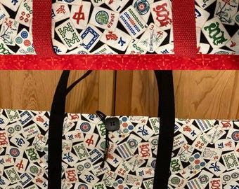 May include: A rectangular fabric tote bag featuring a repeating pattern of Mahjong tiles on a black background. The bag includes black fabric handles and a single black button closure with a cord loop. The top section shows red fabric trim.
