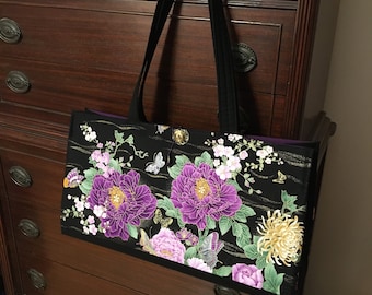 Op de afbeelding: Een zwarte tote bag met een levendig bloemenpatroon van grote paarse pioenrozen, gele chrysanten, groene bladeren en kleine vlinders. De tas heeft twee zwarte schouderbanden en een goudkleurige knoopsluiting met een koordlus.