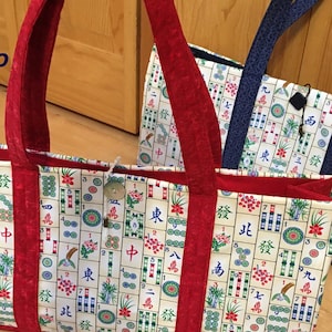 Op de afbeelding: Twee tote tassen met een crèmekleurige achtergrond en een patroon van Mahjong tegels. De tassen hebben rode en blauwe handvatten en afwerking. Eén tas heeft een houten knoopsluiting.