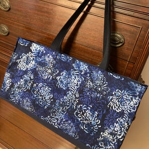 May include: A rectangular tote bag with a dark blue base and straps. The bag's main body features a floral pattern in shades of blue and white. The bag is sitting on a wooden dresser.