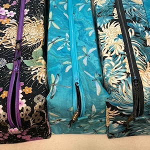 Può includere: Cinque astucci rettangolari in tessuto con cerniera disposti fianco a fianco. Gli astucci presentano vari motivi floreali e di libellule in tonalità di blu, nero, viola e verde acqua, ciascuno dotato di una chiusura a cerniera coordinata.