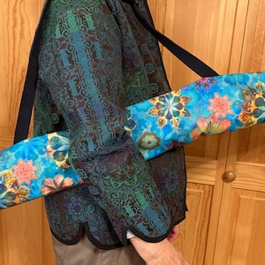 May include: A blue yoga mat bag with a floral pattern in shades of orange, yellow, and green. The bag has a black shoulder strap and a drawstring closure. The bag is designed to carry a standard-sized yoga mat.