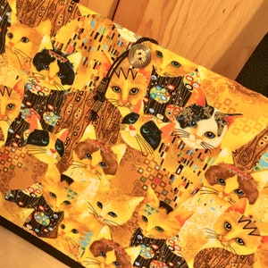 May include: A tote bag featuring a repeating pattern of cat faces in a style reminiscent of Gustav Klimt's artwork. The cats have various fur colors, including orange, black, and white, set against a gold background. The bag has a black strap and a button closure.