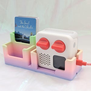May include: A white audio player with red controls, set in a gradient tray. The tray, in shades of pink, green, and purple, holds cards with "The Snail and the Whale" text. A red and white cable is connected to the device.