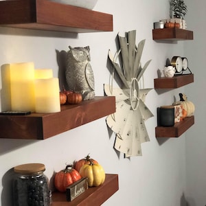 May include: A display of decorative items on floating shelves. Includes a concrete bowl, an owl figurine, candles, pumpkins, a jar of black beans, and a windmill decoration. The shelves are stained a dark brown.
