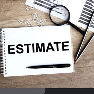 May include: A white spiral-bound notebook with the word "ESTIMATE" in bold black letters. A black pen rests on the notebook. A magnifying glass, pencils, paperclips, and charts are also on the wooden surface.
