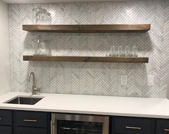 Op de afbeelding: Twee houten planken gemonteerd op een witte visgraat betegelde muur boven een wit aanrecht met een roestvrijstalen gootsteen en een wijnkoeler. De planken zijn donkerbruin gebeitst en leeg, behalve een glazen pot op de bovenste plank en een rij heldere glazen op de onderste plank.