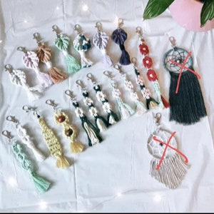 May include: A collection of colorful macrame keychains with various designs, including flowers, dreamcatchers, and geometric patterns. The keychains are made with natural fibers and have tassels.