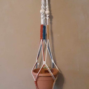 May include: Macramé plant hanger with a terracotta pot. The hanger features a white hook, red, tan, blue, and white knotted cords, and wooden beads. The pot is suspended by the macramé, creating a decorative display.