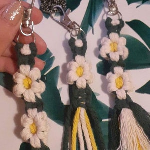 May include: Three green, white, and yellow macrame keychains with white daisy flowers. Each keychain has a silver key ring.
