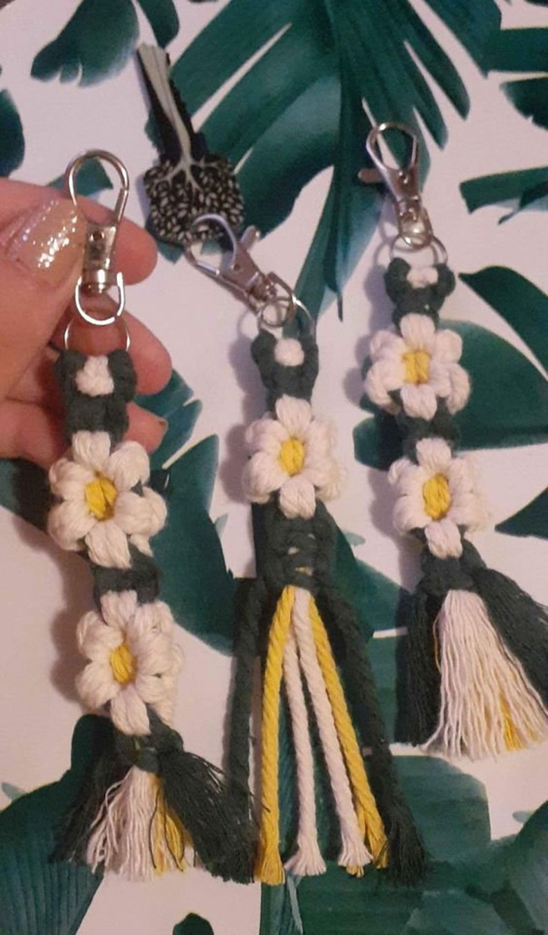 May include: Three green, white, and yellow macrame keychains with white daisy flowers. Each keychain has a silver key ring.