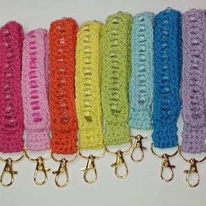May include: Ten crocheted keychains with colorful beads and gold key rings. The keychains are in the colors white, pink, red, orange, yellow, green, light blue, dark blue, purple, and dark blue.