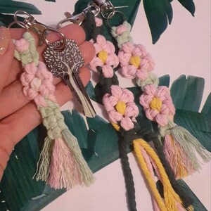 May include: Three handmade macrame keychains with pink, green, and yellow tassels. Each keychain features a flower design with pink and yellow accents.