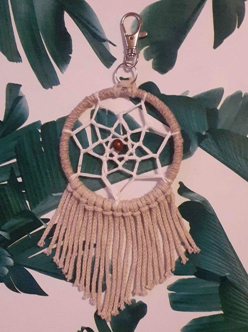 May include: A beige dreamcatcher keychain with a white web and a brown bead in the center. The keychain has a silver clip.