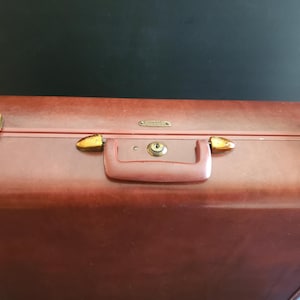 May include: A rectangular brown suitcase featuring a central handle with a built-in key lock mechanism. The luggage includes two metallic side latches and a small rectangular brand plate centered above the handle on the top edge.
