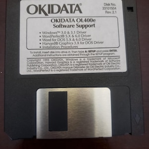 May include: A black floppy disk with a silver label. The label reads "OKIDATA OL400e Software Support" and lists the software drivers included: Windows 3.0 & 3.1, WordPerfect 5.X & 6.0, Word for DOS 5.X & 6.0, Harvard Graphics 3.X for DOS, and Installation Procedures. The disk number is 33101504 and the revision is 2.1.