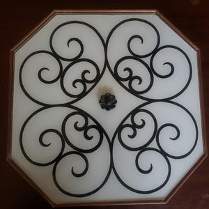 May include: An octagonal ceiling light fixture with a frosted glass center. The fixture features a black wrought iron design of swirling patterns and a small black flower-shaped center detail. The frame is a warm copper color.