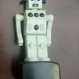 May include: A light green plastic robot toy with black eyes, a black hat, and black details. The robot is standing on a black base.