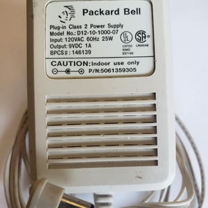 May include: A beige Packard Bell Plug-in Class 2 Power Supply with a white cord. The power supply has the model number D12-10-1000-07 and includes the text: Input: 120VAC 60Hz 25W, Output: 9VDC 1A, and CAUTION: Indoor use only.