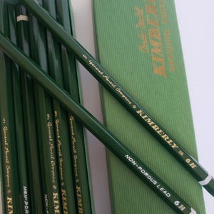 May include: A set of dark green Kimberly 6H drawing pencils by General Pencil Company, featuring gold lettering and white end caps. The pencils are displayed alongside a matching green cardboard box with gold text reading Kimberly Drawing Pencils.