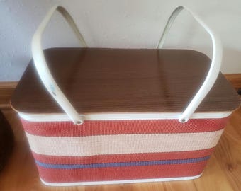 May include: A vintage picnic basket with a brown wooden lid and white handles. The basket's body features horizontal stripes in red, white, and blue. The basket is designed for carrying food and drinks for outdoor dining.