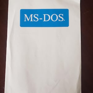 May include: A white paper with a blue rectangular label at the top center. The label contains the text "MS-DOS" in white, sans-serif font. The paper is slightly crumpled.