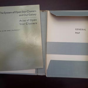 May include: An open book with a light green cover titled "The System of Open Star Clusters and Our Galaxy" and "Atlas of Open Star Clusters." The book is open to a page with the text "GENERAL MAP."