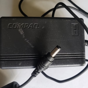 May include: Black Compaq power adapter with a DC connector and a power cord. The adapter has the brand name "COMPAQ" printed on the top. The power cord is black.