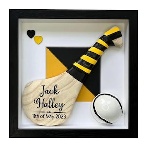 Puede incluir: Obra de arte enmarcada con un stick de hurling de madera, empuñadura negra y amarilla, y una pelota blanca y negra. El stick lleva la inscripción "Jack Halley" y la fecha "11 de mayo de 2023". Dos corazones completan la composición.