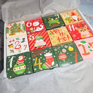 May include: A white box containing twelve square cardboard gift boxes arranged in a grid, each featuring unique Christmas-themed illustrations and numbers from one to twelve, including images of Santa, reindeer, snowmen, and festive ornaments.