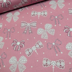May include: A dusty rose fabric featuring a repeating pattern of bows rendered in white and light pink dots. The design includes various bow shapes, some with dangling beaded strands, interspersed with small white polka dots across the surface.