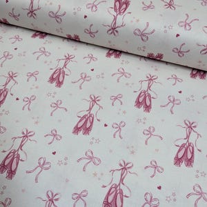 May include: A white fabric featuring a repeating pattern of dark pink ballet slippers, ribbons, small hearts, and stars. The design is printed across the surface of the textile, showcasing a consistent motif of dance-themed elements.