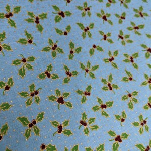 May include: A light blue fabric with a repeating pattern of green holly leaves and red berries. The background is a light blue with a subtle gold glitter pattern.
