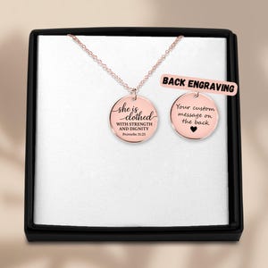 Peut inclure: Un collier en or rose avec un pendentif rond, présenté dans un coffret noir. Le pendentif porte l'inscription "she is clothed with strength and dignity Proverbs 31:25". Un second pendentif affiche "Your custom message on the back".