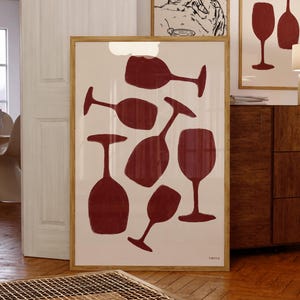 May include: A framed print featuring a collection of stylized wine glass silhouettes in a deep red hue against a cream background. The artwork is displayed in a room with other framed art and wooden furniture.