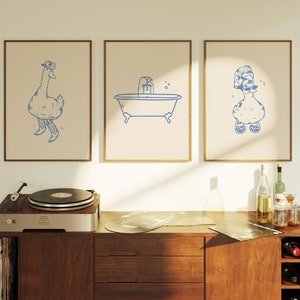 May include: Three framed illustrations on a white wall above a wooden cabinet. The illustrations are of a duck wearing a cowboy hat, a bathtub with a tap, and a duck wearing a shower cap.