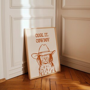 Op de afbeelding: Een ingelijst print met een bruine rand toont een lijntekening van een hond die een cowboyhoed draagt. De tekst "COOL IT, COWBOY" is in bruin boven de hond gedrukt.