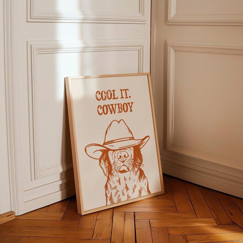 Op de afbeelding: Een ingelijst print met een bruine rand toont een lijntekening van een hond die een cowboyhoed draagt. De tekst "COOL IT, COWBOY" is in bruin boven de hond gedrukt.