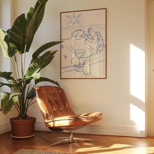 May include: A framed art print featuring a dog relaxing with a cocktail on a beach, rendered in blue line art. The print hangs above a brown leather chair with a metal base, next to a large potted plant.