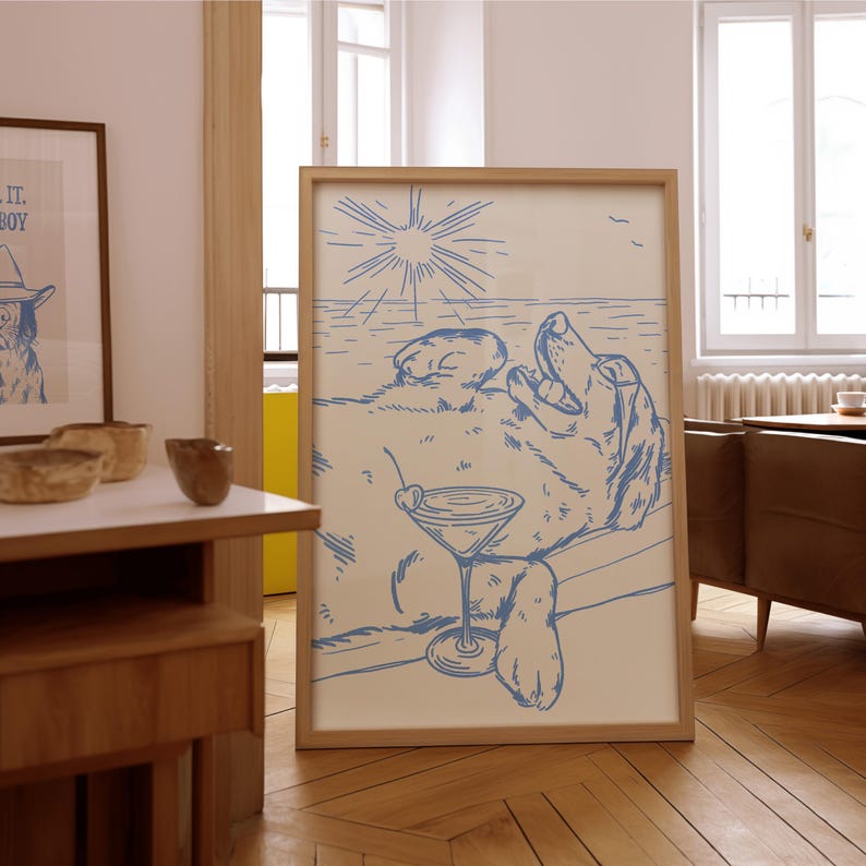 May include: A framed print featuring a dog relaxing on a beach, holding a cocktail. The artwork is in a blue line drawing style, with a sun and ocean in the background. The frame is made of light-colored wood.