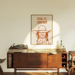Op de afbeelding: Een bruin houten kast met een platenspeler erop. De kast heeft een platenrek aan de linkerkant en een wijnrek aan de rechterkant. Een ingelijst afdruk met een hond die een cowboyhoed draagt en de tekst "COOL IT. COWBOY" hangt aan de muur achter de kast.