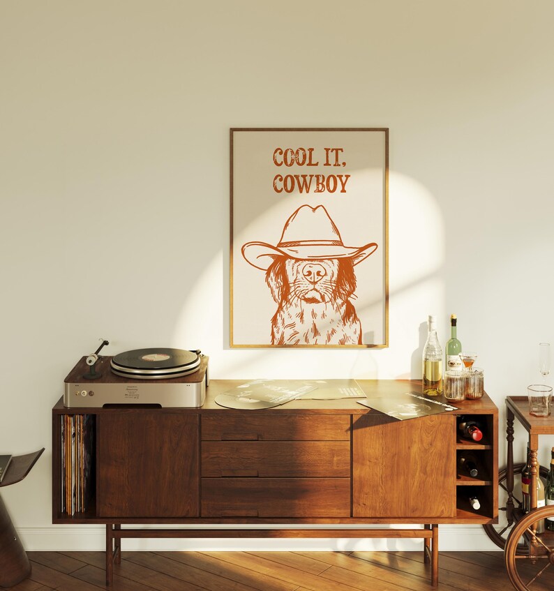 Op de afbeelding: Een bruin houten kast met een platenspeler erop. De kast heeft een platenrek aan de linkerkant en een wijnrek aan de rechterkant. Een ingelijst afdruk met een hond die een cowboyhoed draagt en de tekst "COOL IT. COWBOY" hangt aan de muur achter de kast.