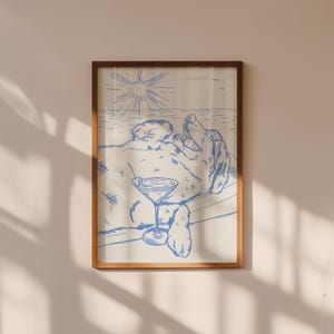 May include: A framed print featuring a blue line drawing of a dog relaxing with a cocktail. The dog is lying on its back, with a martini glass in its paw. The background depicts a beach scene with the sun and ocean.