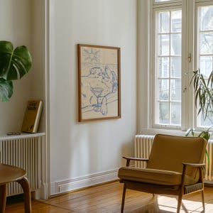 May include: A framed artwork depicts a person relaxing with a cocktail under a sun. The artwork is in a wooden frame and hangs on a white wall. A mustard-colored armchair sits on a wooden floor, next to a window.