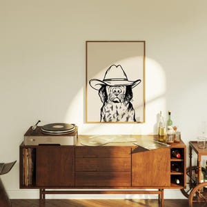 May include: A black and white line drawing of a dog wearing a cowboy hat. The drawing is framed and hanging on a wall above a wooden cabinet with a record player on top.