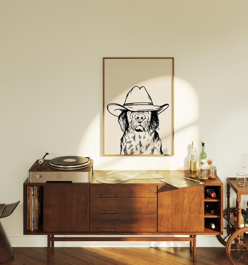 May include: A black and white line drawing of a dog wearing a cowboy hat. The drawing is framed and hanging on a wall above a wooden cabinet with a record player on top.