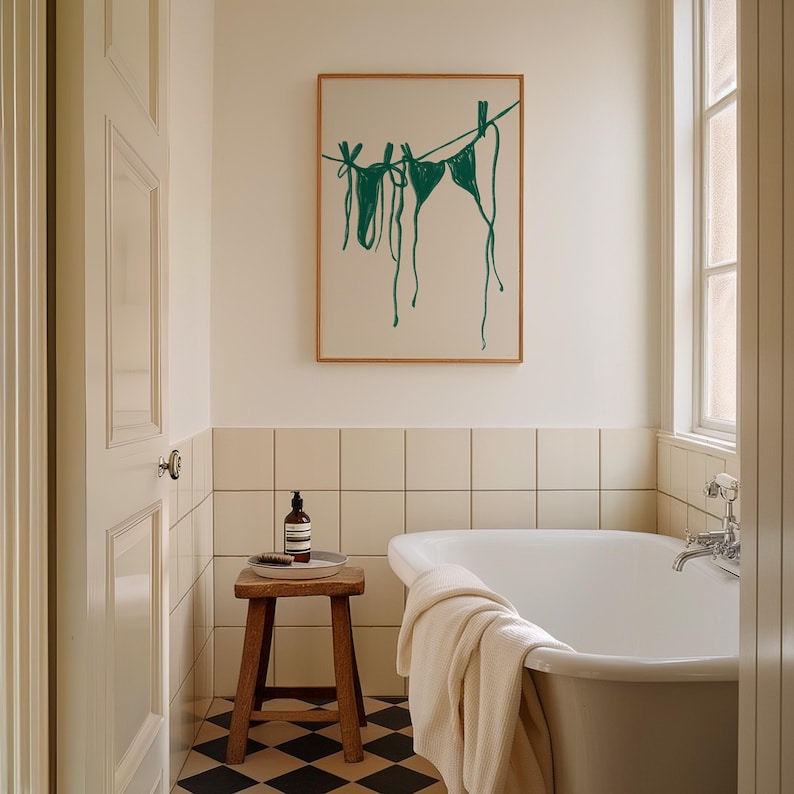 May include: A white bathroom with a bathtub, a wooden stool, and a framed print of green bikini tops hanging on a clothesline.